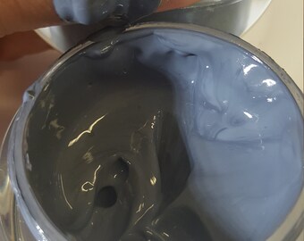Op de afbeelding: Een close-up van een grijs houtskool gezichtsmasker in een zilveren pot. Het masker is dik en romig en heeft een gladde, gelijkmatige textuur.