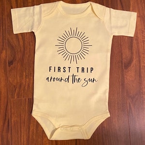 Puede incluir: Un body de bebé amarillo con un gráfico de sol negro y el texto "FIRST TRIP around the sun".