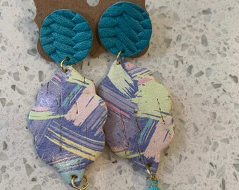 Peut inclure: Une paire de boucles d'oreilles avec un cercle en cuir bleu sarcelle et un motif abstrait coloré avec un pompon bleu sarcelle.