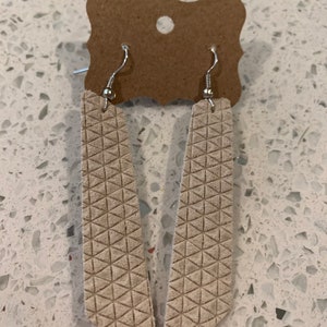 Puede incluir: Un par de aretes largos de cuero blanco con un patrón geométrico. Los aretes tienen ganchos plateados.