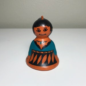 Puede incluir: Figura de campana de cerámica pintada a mano que representa a una persona con vestimenta tradicional. La campana presenta un peinado negro, una parte superior turquesa y una falda naranja con diseños geométricos negros y naranjas. Una pequeña cuerda marrón está unida a la parte superior.