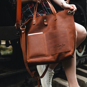 May include: A brown leather tote bag with a long strap and a smaller pocket on the front. The bag has a floral design and the initials "AB" engraved on the front.