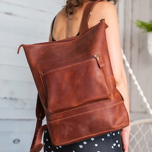 May include: A brown leather backpack with a zippered front pocket. The backpack is worn by a person with a black and white polka dot dress.