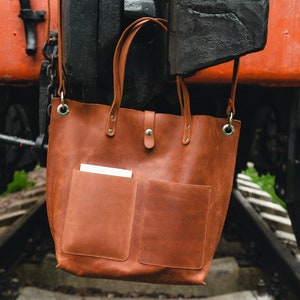 May include: A brown leather tote bag with two front pockets and a white piece of paper in the left pocket. The bag is hanging from a metal hook on a rusty train car.