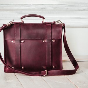 May include: A burgundy leather briefcase with a top handle and an adjustable shoulder strap. The bag has a flap closure with metal buckles and studs.