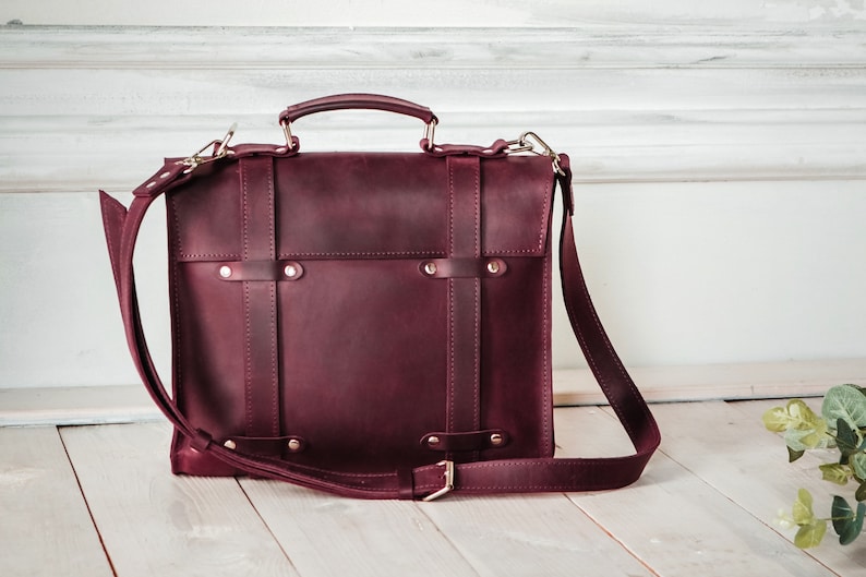 May include: A burgundy leather briefcase with a top handle and an adjustable shoulder strap. The bag has a flap closure with metal buckles and studs.