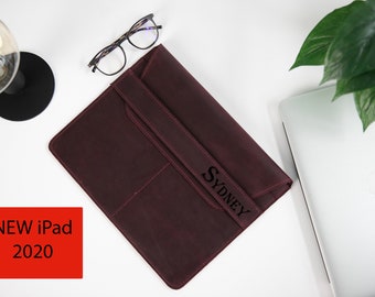 May include: A burgundy leather iPad case with a flap closure and a personalized name "SYDNEY" engraved on the front. The case is on a white surface with a laptop and a plant in the background.