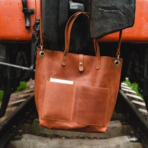 May include: A brown leather tote bag with two front pockets hangs from a rusty metal structure. The bag has a top zipper closure and two metal rings on the sides.