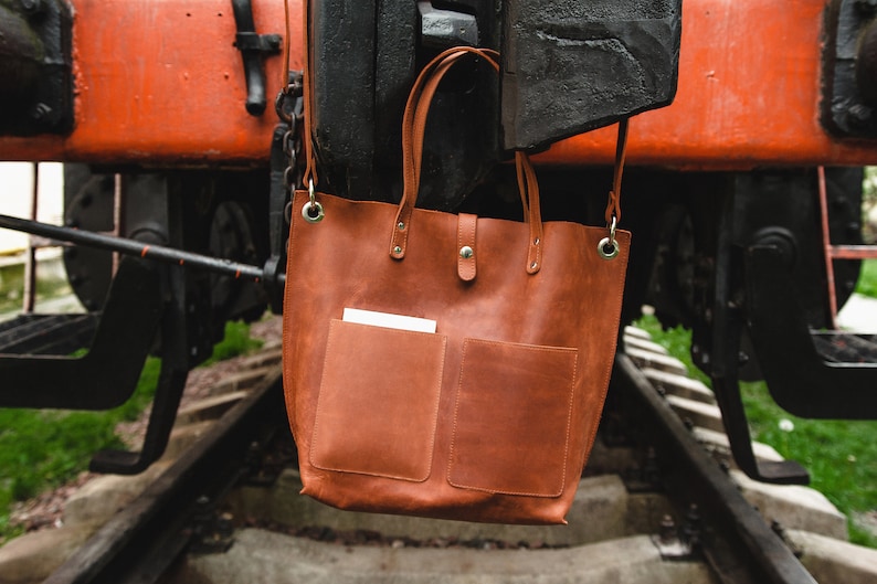 May include: A brown leather tote bag with two front pockets hangs from a rusty metal structure. The bag has a top zipper closure and two metal rings on the sides.