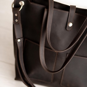 May include: Dark brown leather tote bag with two exterior pockets and a long, adjustable shoulder strap. The bag features a top handle and is accented with silver-tone hardware and visible stitching.