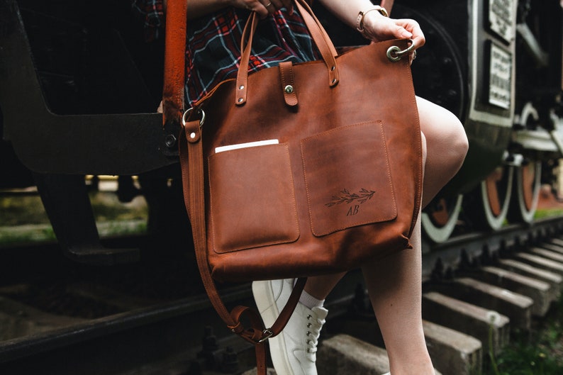 May include: A brown leather tote bag with a long strap and a smaller pocket on the front. The bag has a floral design and the initials "AB" engraved on the front.