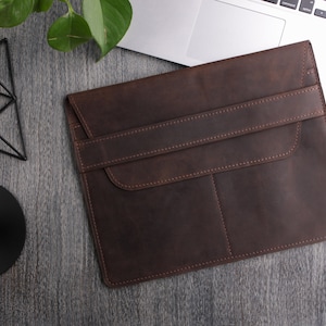 May include: A brown leather laptop sleeve with a flap closure and a pocket on the front. The sleeve is lying on a gray wooden surface.