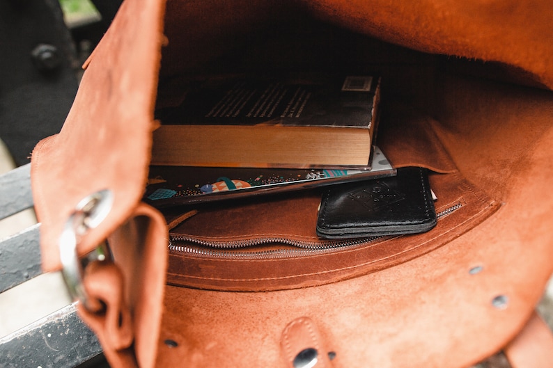 May include: The inside of a brown leather bag with a zippered pocket. A book and a black wallet are inside the bag.