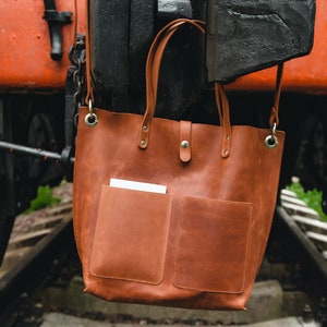 May include: A brown leather tote bag with two front pockets and a long strap. The bag is hanging from a metal hook on a train.