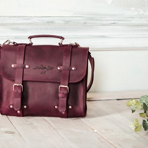 May include: A burgundy leather satchel with a long strap and two buckles. The bag has a handle and is embossed with the initials "AB" and a floral design.