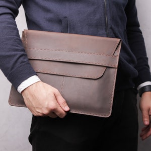 May include: A brown leather laptop sleeve with a flap closure and a strap detail. The sleeve is held by a person wearing a navy blue jumper and a white shirt. The leather has a textured appearance.