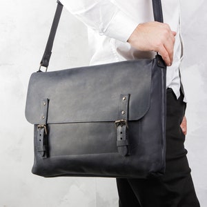 May include: A dark grey leather messenger bag with two buckles and a flap closure. The bag is worn over the shoulder of a person wearing a white shirt and black trousers.