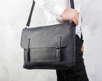 Könnte beinhalten: Eine dunkelgraue Ledertasche mit zwei Schnallen und einem Klappverschluss. Die Tasche wird über der Schulter einer Person getragen, die ein weißes Hemd und eine schwarze Hose trägt.