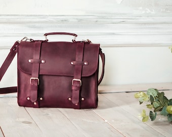 May include: A burgundy leather briefcase with two buckles and a shoulder strap. The bag is sitting on a light wood surface.
