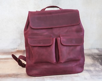 May include: A burgundy leather backpack with a flap closure and two front pockets. The bag has a top handle and adjustable shoulder straps. The leather has a slightly distressed appearance.