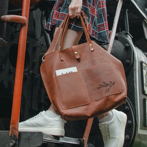 May include: A brown leather tote bag with a large pocket on the front. The pocket has a floral design and the initials "AB" engraved on it.