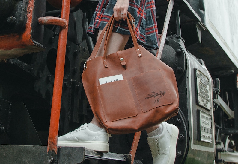May include: A brown leather tote bag with a large pocket on the front. The pocket has a floral design and the initials "AB" engraved on it.