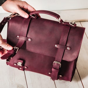 May include: A burgundy leather briefcase with a top handle and adjustable shoulder strap. The bag has multiple buckles and metal accents.