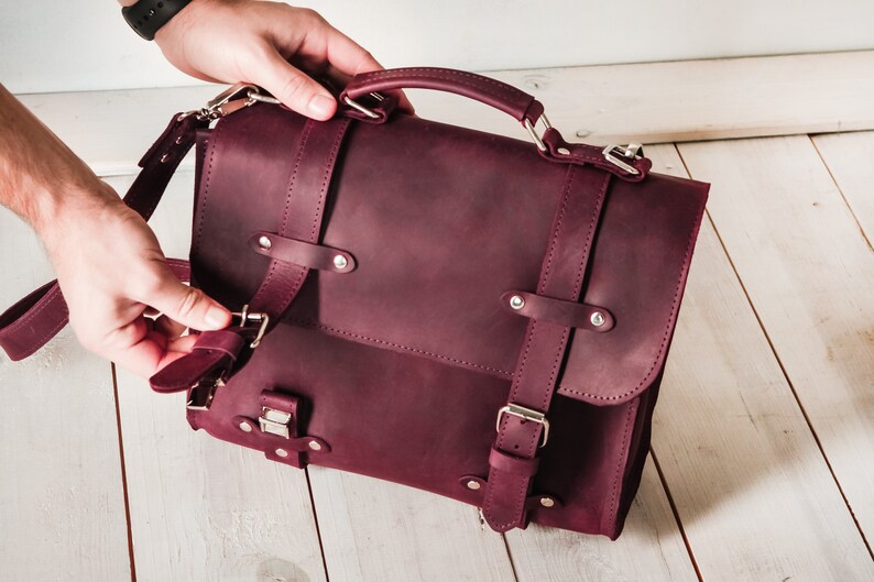 May include: A burgundy leather briefcase with a top handle and adjustable shoulder strap. The bag has multiple buckles and metal accents.