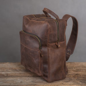 Op de afbeelding: Een bruine leren rugzak met een ritsvak aan de voorkant en verstelbare schouderbanden.