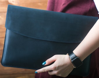 May include: A dark blue leather laptop sleeve with a flap closure. The sleeve is held in a person's hand.