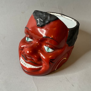 Peut inclure: Un pot en céramique en forme de visage noir souriant avec des yeux verts. Le pot est rouge avec des cheveux noirs et un intérieur blanc.