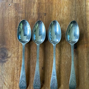 Könnte beinhalten: Ein Set aus vier silberfarbenen Metalllöffeln, die in einer Reihe angeordnet sind. Jeder Löffel hat eine ovale Laffe und einen schlanken Stiel, der mit einem sich wiederholenden Kreuzschraffur-Randmuster und einem kleinen Monogramm verziert ist.