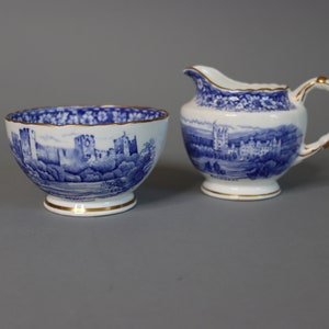 Puede incluir: Un juego de azucarero y jarra de crema de porcelana azul y blanca con ribete dorado. El juego presenta una ilustración azul y blanca de un castillo y el paisaje circundante.