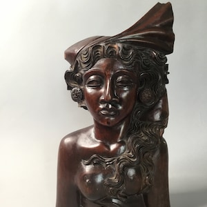 May include: A dark brown wooden sculpture of a woman's bust with intricate details in the hair and clothing. The sculpture is carved in a realistic style and features a serene expression on the woman's face.