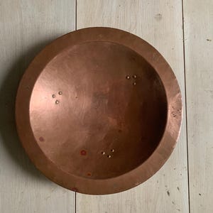 May include: A circular copper bowl with a wide, flat rim and a smooth, metallic finish. The interior surface features three distinct clusters of small, evenly spaced rivets arranged in a triangular pattern, adding texture to the warm-toned metal.