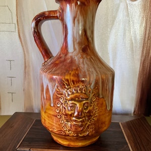 Könnte beinhalten: Eine braune und weiße Keramikkanne mit einem Reliefgesicht auf der Vorderseite. Die Kanne hat eine weiße Glasur, die an den Seiten herunterläuft.