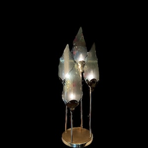 May include: A brass table lamp with three leaf-shaped glass shades. The shades are translucent and have a delicate pattern. The lamp is lit and the light shines through the glass.