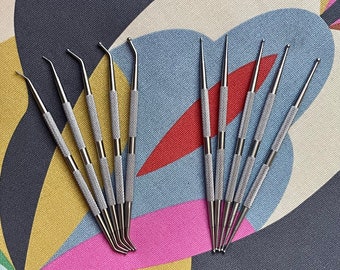 Puede incluir: Un conjunto de 12 herramientas de metal plateado con puntas de diferentes formas. Las herramientas están dispuestas en forma de abanico sobre una tela estampada de colores.
