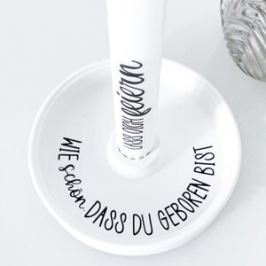 May include: White ceramic stand with a round base and a tall, cylindrical post. The base has the text "WIE SCH&Ouml;N, DASS DU GEBOREN BIST" written around the edge. The post has the text "DASS DU GEBOREN" written on it.
