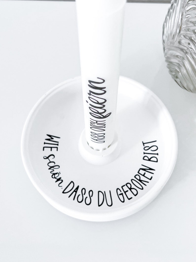 May include: White ceramic stand with a round base and a tall, cylindrical post. The base has the text "WIE SCH&Ouml;N, DASS DU GEBOREN BIST" written around the edge. The post has the text "DASS DU GEBOREN" written on it.