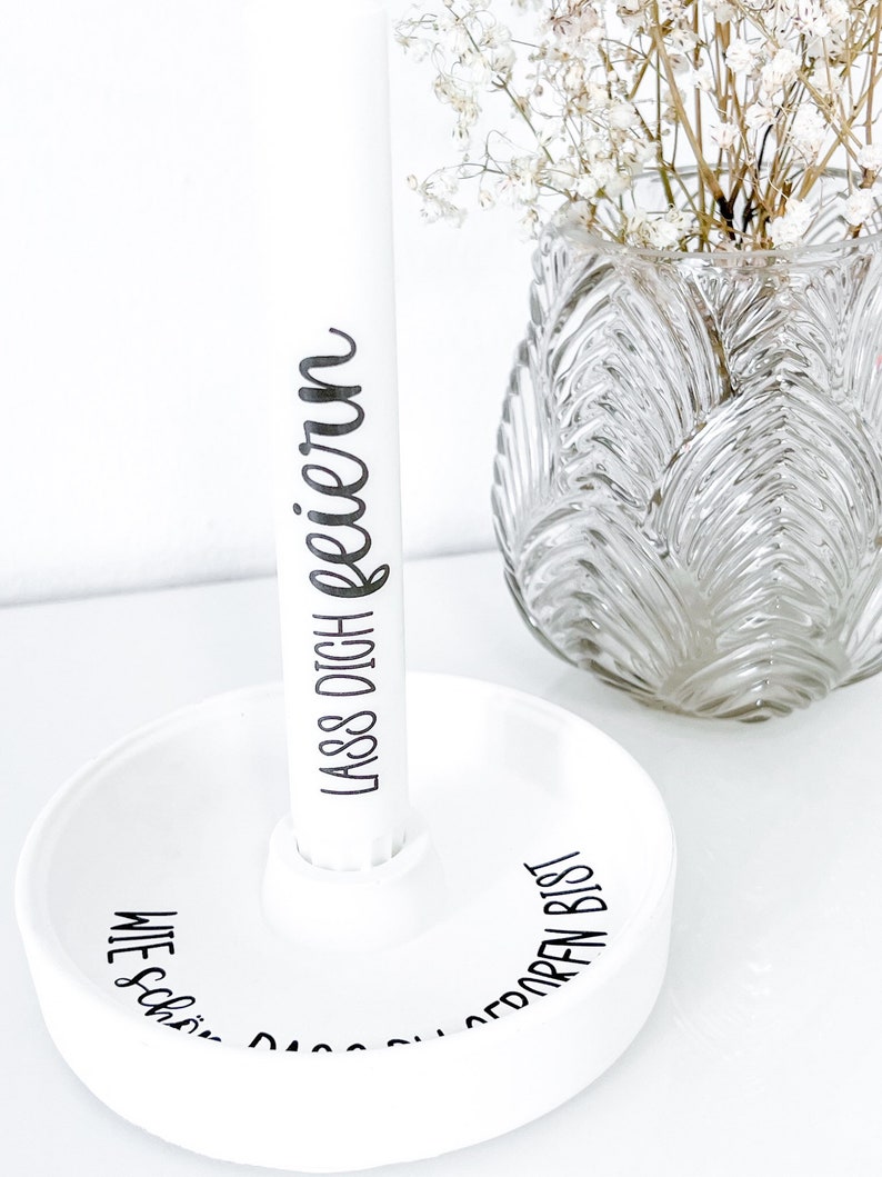 May include: White ceramic holder with the text "Wie sch&ouml;n, dass du bist" and "Lass dich feiern" on the side. The holder is holding a white cylinder with the text "Lass dich feiern" on it.