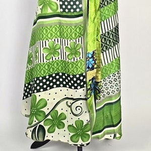 Può includere: Una gonna a portafoglio patchwork nei toni del verde, bianco e nero, esposta su un manichino nero. La gonna presenta motivi floreali, a pois e geometrici. La gonna è legata in vita con una fascia verde abbinata.
