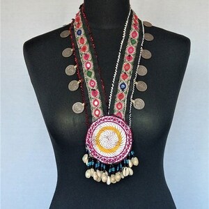 May include: A multi-strand necklace with a large, round, embroidered pendant. The necklace is made of silver coins, beads, and colorful fabric. The pendant is white with a pink and yellow swirl design.