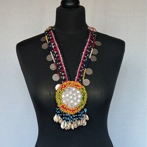 May include: A colorful beaded necklace with a large circular pendant featuring a white center and a multi-colored border. The necklace also includes silver coins and white shell accents.