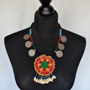 Op de afbeelding: Een kleurrijke kralenketting met een grote, ronde hanger met een stervormig ontwerp in tinten rood, oranje, groen en blauw. De hanger is omringd door zilveren munten en de ketting is afgewerkt met een franje van witte schelpen.