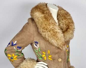May include: A tan suede coat with a faux fur collar and cuffs. The coat features embroidered floral designs in yellow, blue, and green on the sleeves. It has wooden buttons and a tailored fit, showcasing a vintage aesthetic.