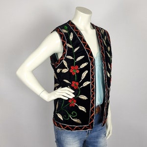 May include: Black velvet vest with floral embroidery. The vest features red flowers, green leaves, and gold and orange accents. The vest is sleeveless and has an open front. The vest is displayed on a mannequin.