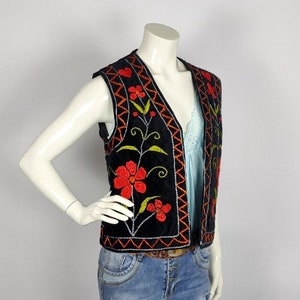 May include: Black velvet vest with red floral embroidery and orange zigzag trim. The vest features a V-neck design and is displayed on a mannequin. The floral design includes red flowers, green leaves, and black stems.