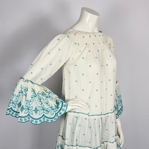 Op de afbeelding: Een witte off-shoulder jurk met een gelaagde rok en wijde mouwen. De jurk is bedekt met kleine blauwe stippen en heeft blauwe geborduurde bloemenmotieven op de mouwen.