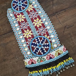 May include: A beaded textile with a vibrant design. The piece features a central pattern of red, white, and yellow floral motifs, bordered by blue beads and small mirrors. Two circular beaded elements are at the top and bottom.